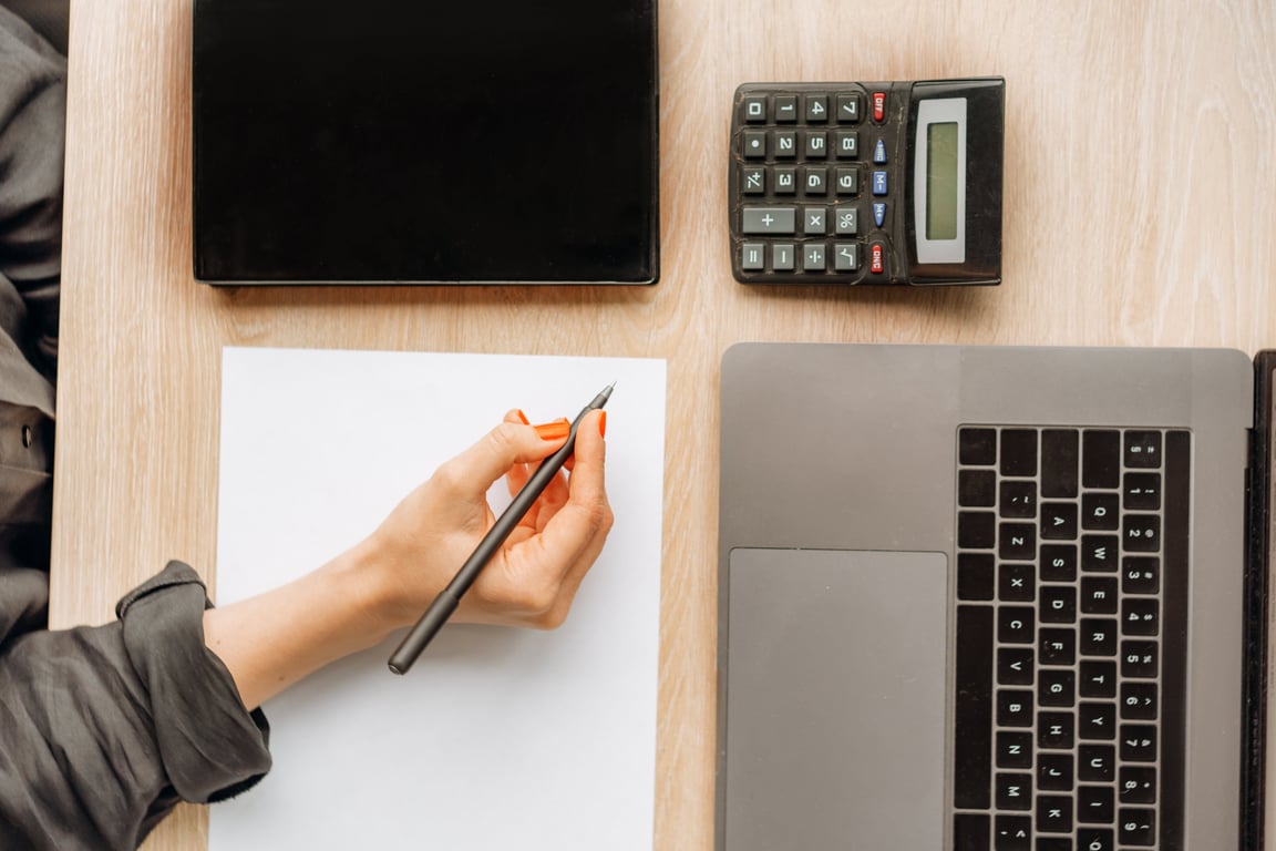 A digital nomad accountant using pen and paper next to their laptop and calculator.