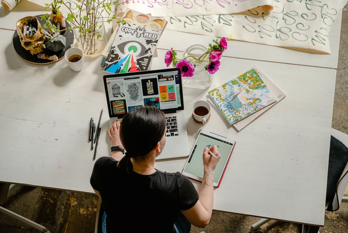 Woman at a desk using a laptop with her left hand and writing notes with her right hand, surrounded by books and drawings.